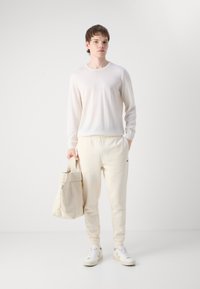 Lichtbeige langemouwen shirt gecombineerd met crèmekleurige sweatpants. Het model houdt een beige tas vast. De outfit heeft een minimalistisch ontwerp en zachte texturen.