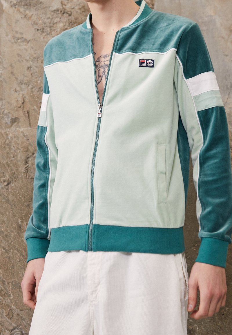 Zamszowa kurtka zapinana na zamek w kolorze mięty z turkusowymi akcentami. Posiada biały pasek i boczne kieszenie. Miękka tekstura, minimalistyczny design.