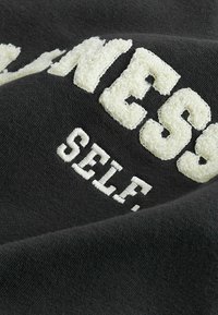 Must värv musta kangast, millel on tekstureeritud valged tikitud tähed "MADNESS SELF." Suurendatud vaade, mis rõhutab pehmet tekstuuri ja kontrastseid värve.