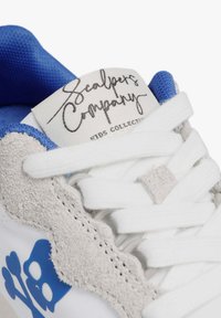 Zapatilla blanca con acentos azules, superposiciones de gamuza y una etiqueta distintiva. Presenta cordones blancos y un diseño decorativo de calavera y huesos cruzados.