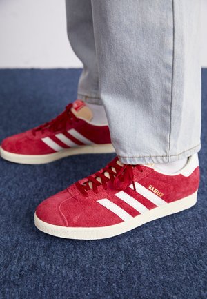 Baskets en daim rouge avec des bandes blanches et un logo doré. Design à lacets et semelle en caoutchouc texturée. Portées avec un pantalon en jean délavé clair.