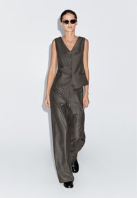 Gray pinstriped vest with a V-neckline and front buttons, paired with loose, wide-leg trousers. Model wears black sunglasses and shoes.