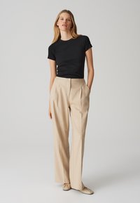 Zwarte aansluitende t-shirt gecombineerd met hoogwassende beige geplooide broeken. De broeken hebben een knoopsluiting en zijzakken, met een gladde textuur.