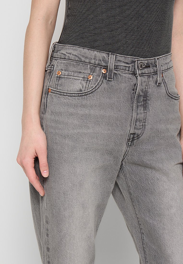Vaqueros de mezclilla gris con un corte de pierna recta, diseño de cinco bolsillos, detalles en hardware de cobre y un acabado desgastado. Se llevan con una blusa negra acanalada.