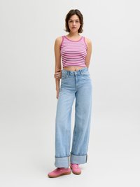 Γυναίκα με ροζ και λευκό ριγέ αμάνικο crop top, φαρδιά ανοιχτό μπλε τζιν με γαζιά και ροζ αθλητικά παπούτσια, όρθια μπροστά σε απλό φόντο.