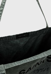 Fekete anyag táska szürke részletekkel, amelynek tetején címke található, és a frontján feltűnő szöveg design látható. Puha tapintású, nyitott tetejű.