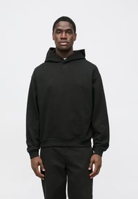 Čierna mikina s kapucňou v uvoľnenom strihu, s rebrovanými manžetami a lemom, s malým logom 'BOSS' na hrudi. Nosená s tmavými nohavicami.