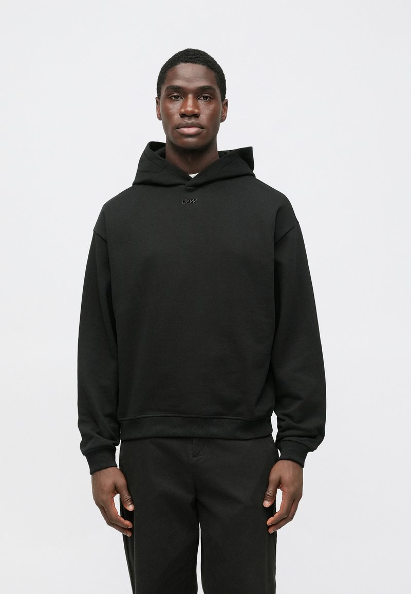 Čierna mikina s kapucňou v uvoľnenom strihu, s rebrovanými manžetami a lemom, s malým logom 'BOSS' na hrudi. Nosená s tmavými nohavicami.