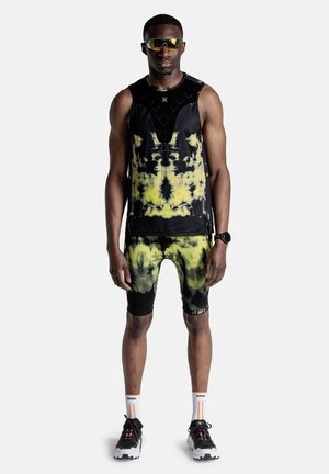 Atleet draagt een zwart en geel tie-dye mouwloos topje en short, zwarte hardloopschoenen, witte sokken, een zonnebril en een fitnesshorloge, staand.