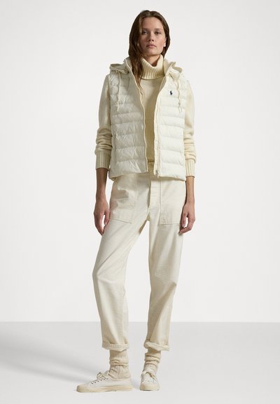 Daunenweste in Off-White mit Kapuze, kombiniert mit einem cremfarbenen Rollkragenpullover und hellbeigen Hosen. Sneaker in einem passenden Farbton mit gerippten Socken.