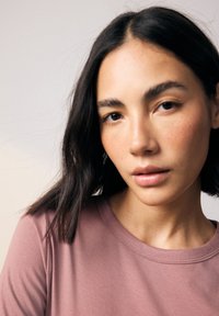 Ljusrosa t-shirt med korta ärmar, rund halsringning och slät textur. Modellen har mörkt, axellångt hår, med en neutral bakgrund.