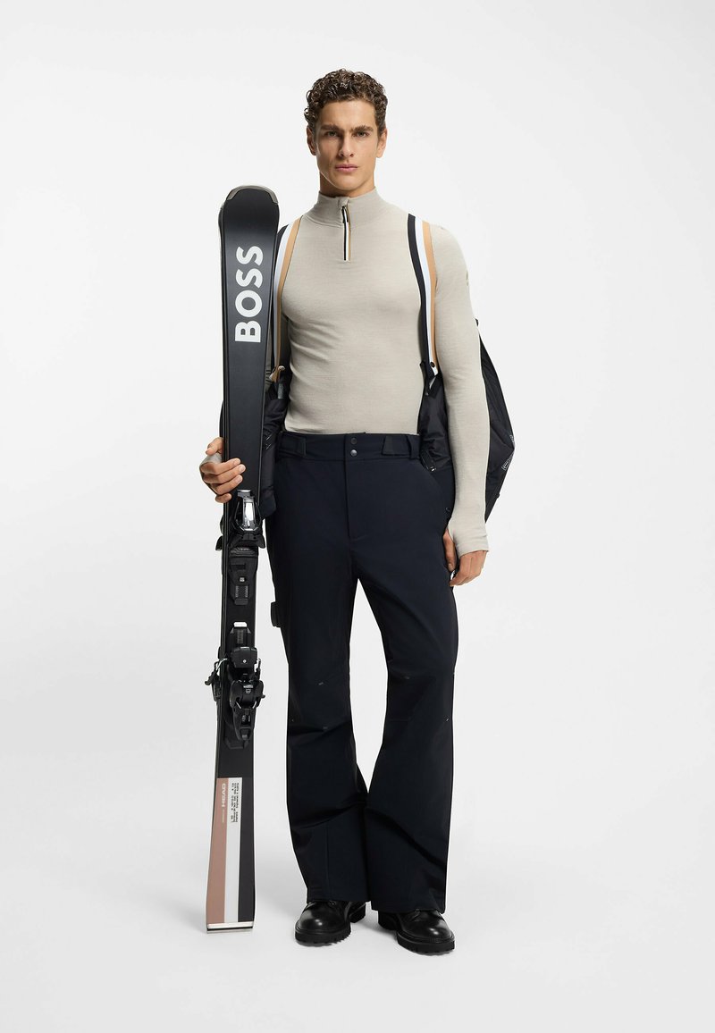 Attrezzatura da sci con un sci nero che mostra "BOSS", abbinato a una maglia beige a maniche lunghe fitted e pantaloni da neve neri, mentre tiene uno zaino.