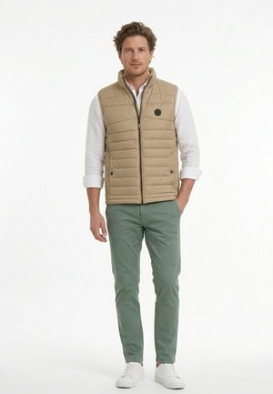 Hombre con chaleco acolchado beige sobre camisa blanca, pantalones verdes y zapatillas blancas, de pie con una mano en el bolsillo.