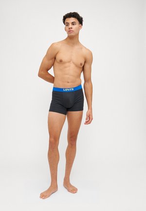 Boxers negros con una cintura azul que presenta un logo. El modelo está descalzo, mostrando el ajuste y diseño del producto.