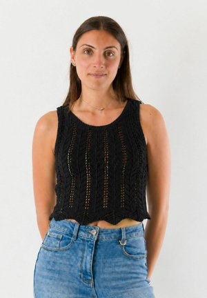 Donna con lunghi capelli castani indossa un top nero lavorato a maglia senza maniche e jeans blu a vita alta, in piedi su sfondo bianco.