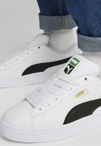 Baskets blanches Puma Suede XL avec détail de logo noir, portées avec des chaussettes blanches et un jean bleu retroussé sur un fond clair.