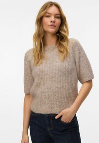 Femme aux cheveux blonds ondulés portant un pull en maille beige à manches courtes et un jean bleu foncé, une main dans la poche, sur un fond blanc uni.