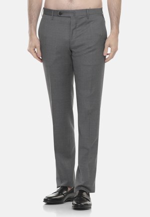 Uomo con pantaloni eleganti grigi e mocassini in pelle nera, in piedi contro uno sfondo bianco uniforme.