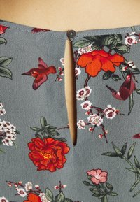 Tela con estampado floral en tonos de gris, rojo y rosa. Presenta una abertura en la parte posterior con llavero asegurada por un botón. Textura suave con patrones detallados.