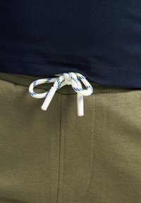 T-shirt en coton bleu marine rentré dans un pantalon de survêtement vert olive. Le pantalon présente un cordon de serrage blanc et vert avec des embouts en plastique. Détail de couture visible.