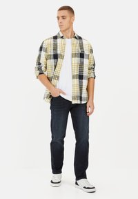 Geruite shirt in geel, zwart en wit, laag over een wit T-shirt; gecombineerd met donkere jeans en witte sneakers.