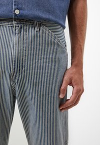 Hombre con pantalones a rayas verticales azules y blancos, camiseta azul metida por dentro, mano derecha relajada al lado.