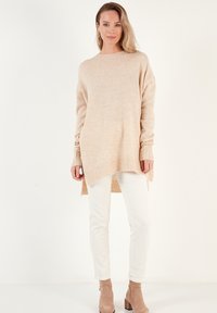 Pull en tricot beige surdimensionné avec des manches longues, des poignets côtelés et un ourlet en pente, associé à un pantalon blanc ajusté et des bottines beige.