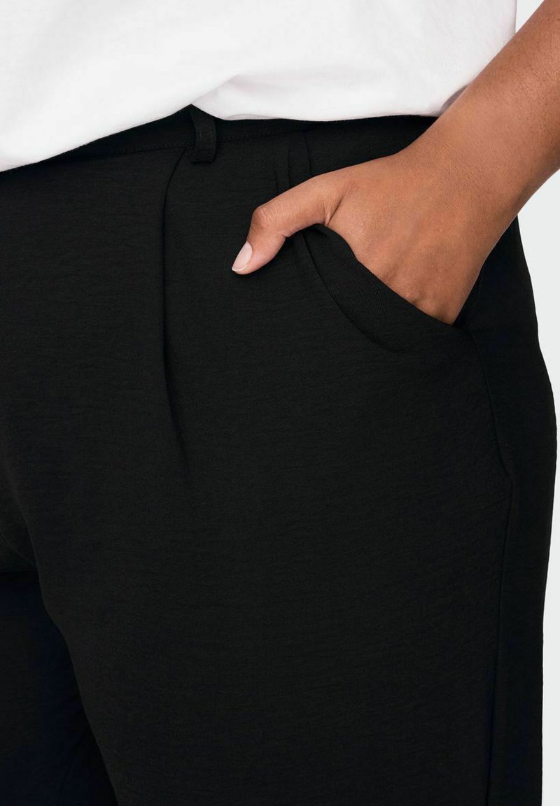 Schwarze Hose aus weichem Stoff, mit Seitentasche, elastischem Bund und subtiler Struktur. Die Hand liegt zur Detailbetonung in der Tasche.