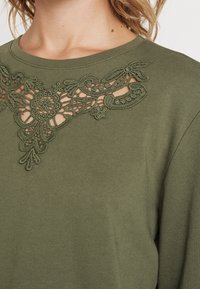 Oliwkowy sweter z detalami koronkowego haftu przy dekolcie, wykonany z teksturowanej tkaniny i w luźnym kroju.