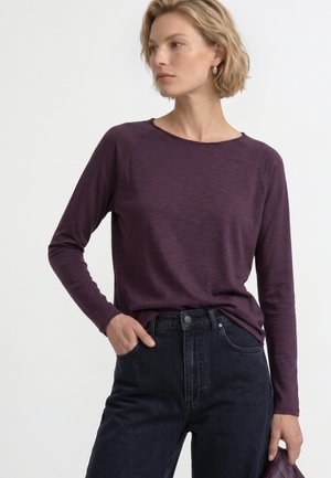 Femeie purtând un top violet cu mâneci lungi și blugi negri cu talie înaltă, pozând cu o mână în buzunar și ținând o geantă clutch violetă.