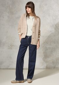 Beige gebreide cardigan met knopen aan de voorkant, gecombineerd met een lichte geribde trui en donkere wijde denim jeans. Neutrale schoenen maken de look af.