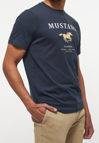 Námorně modré bavlněné tričko s grafikou světle béžového koně a textem "MUSTANG CLASSICS" přes hruď, kulatý výstřih, krátké rukávy.