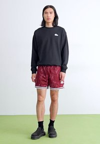 Schwarzer Sweatshirt, kastanienbraune gemusterte Shorts mit weißen Akzenten, schwarze Socken und Slipper, stehend auf grünem Boden vor weißem Hintergrund.