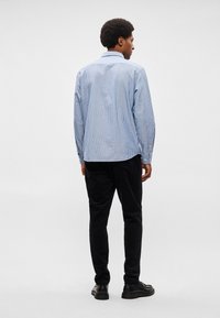 Ανδρική μπλούζα με μπλε και λευκές ρίγες, μακριά μανίκια, και χαρακτηριστικό γιακά, με κουμπιά στους μανίκιες και καμπυλωτό στρίφωμα, συνδυασμένη με μαύρες κοτλέ παντελόνες.