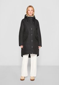 Juodas vidutinio ilgio vandeniui atsparus paltas su gobtuvu, priekiniais sagčių užsegimais ir dviem priekinėmis kišenėmis. Lygus paviršius ir laisvas siluetas.