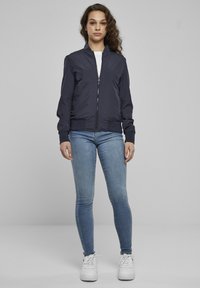 Jachetă bomber bleumarin cu fermoar frontal, manșete și tiv striate. Poartă-o peste un tricou alb și asortează cu blugi bleu deschis și adidași albi.