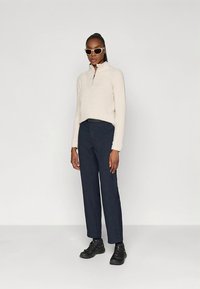 Giacca in pile bianca con cerniera, pantaloni blu navy e scarpe nere. Modello con occhiali da sole oversize, in piedi con le mani lungo i fianchi su uno sfondo semplice.