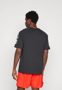 Čierne bavlnené tričko s krátkymi rukávmi, vybavené okrúhlym výstrihom a bielym grafickým detailom na pravom rukáve. Nosí sa s červenými šortkami.