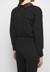 Černá mikina s přiléhavým střihem, s kontrastním černým koženým límcem a žebrovanými manžetami. Měkká textura a elastický pas.