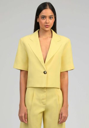 Femme aux longs cheveux foncés porte un blazer jaune pâle à manches courtes avec un bouton et un pantalon taille haute assorti.