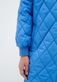 Chaqueta azul acolchada con puños acanalados, que presenta un patrón de diamantes y una textura suave, mostrando parte de una mano contra un fondo claro.