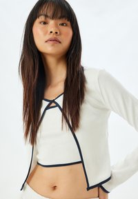 Witte geribbelde crop top met lange mouwen, voorzien van een marineblauwe rand en een open voorkant, vastgemaakt met een knoop bij de hals.