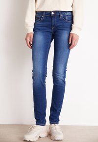 Skinny blue denim jeans with a faded texture, five-pocket design, and contrasting stitching, paired with beige athletic shoes.