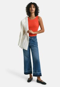 Rode geribbelde tanktop, witte gestreepte blazer, wijde denim jeans met een vervaagde zoom en zwarte platte schoenen. Krullend haar en neutrale expressie.