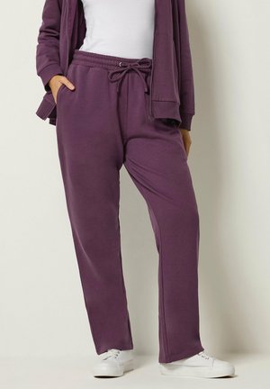 Person trägt violette Jogginghose mit Kordelzug, passende Jacke, weißes Hemd und weiße Sneakers und steht vor einer einfachen Wand.