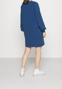 Blaues Sweatshirtkleid mit langen Ärmeln, rundem Ausschnitt und lockerer Passform, kombiniert mit weißen High-Top-Sneakern. Glatte Textur, keine Muster.