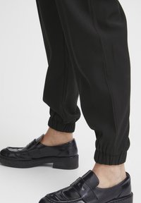 Zwarte jogpants met elastische boorden, gecombineerd met glanzende zwarte instappers met een band over de wreef en een lage, stevige hak.