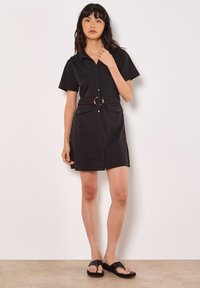 Robe noire à manches courtes avec un devant à boutons, deux poches et une ceinture à la taille avec une boucle en or ronde. Texture de tissu lisse.
