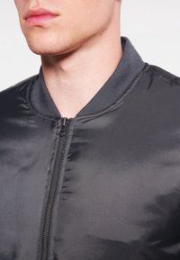 Veste bomber noire avec une texture lisse et brillante, dotée d'un col côtelé et d'une fermeture éclair à l'avant. Aucun motif visible.
