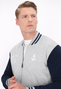 Jachetă bomber gri și bleumarin cu fermoar în față, manșete cu dungi și un logo cu ancoră. Caracteristici: material neted cu mâneci contrastante.